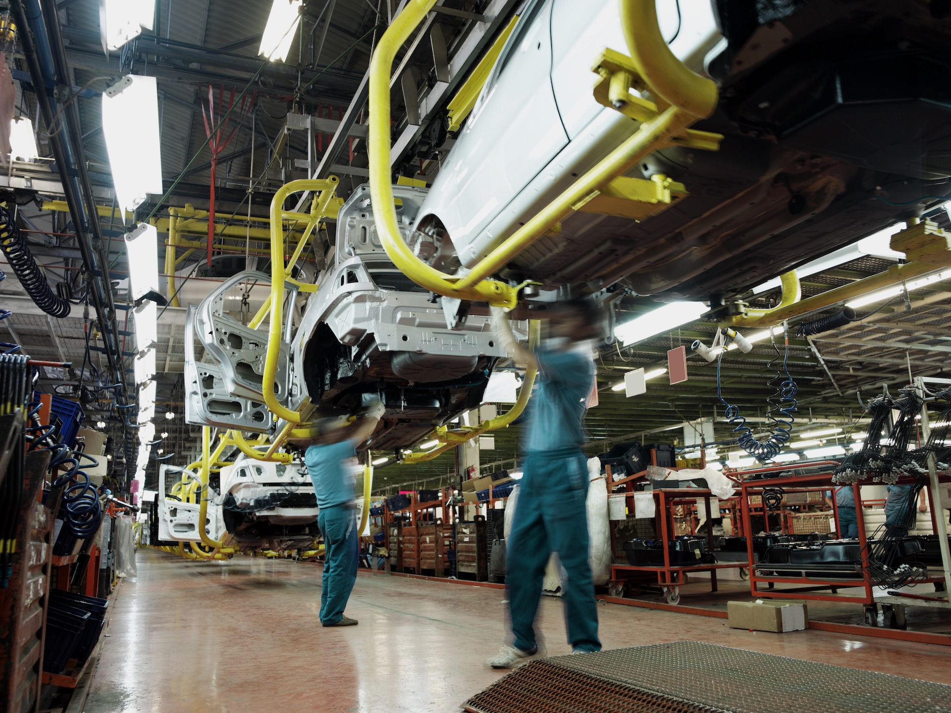 Two workers wearing green shirts and blue pants work underneath a car in an automobile factory. There are assembly lines, partially constructed cars, tools and electrical equipment in the background.